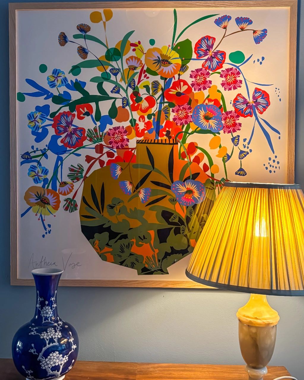 Antheia Vase fine art print by Kitty McCall in a natural oak frame, styled on a sideboard beside a blue-and-white ceramic vase and a yellow pleated lamp