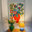 Red Vase fine art print by Kitty McCall in a white frame, styled in a home corner with a green ceramic vase of yellow tulips and a yellow table lamp