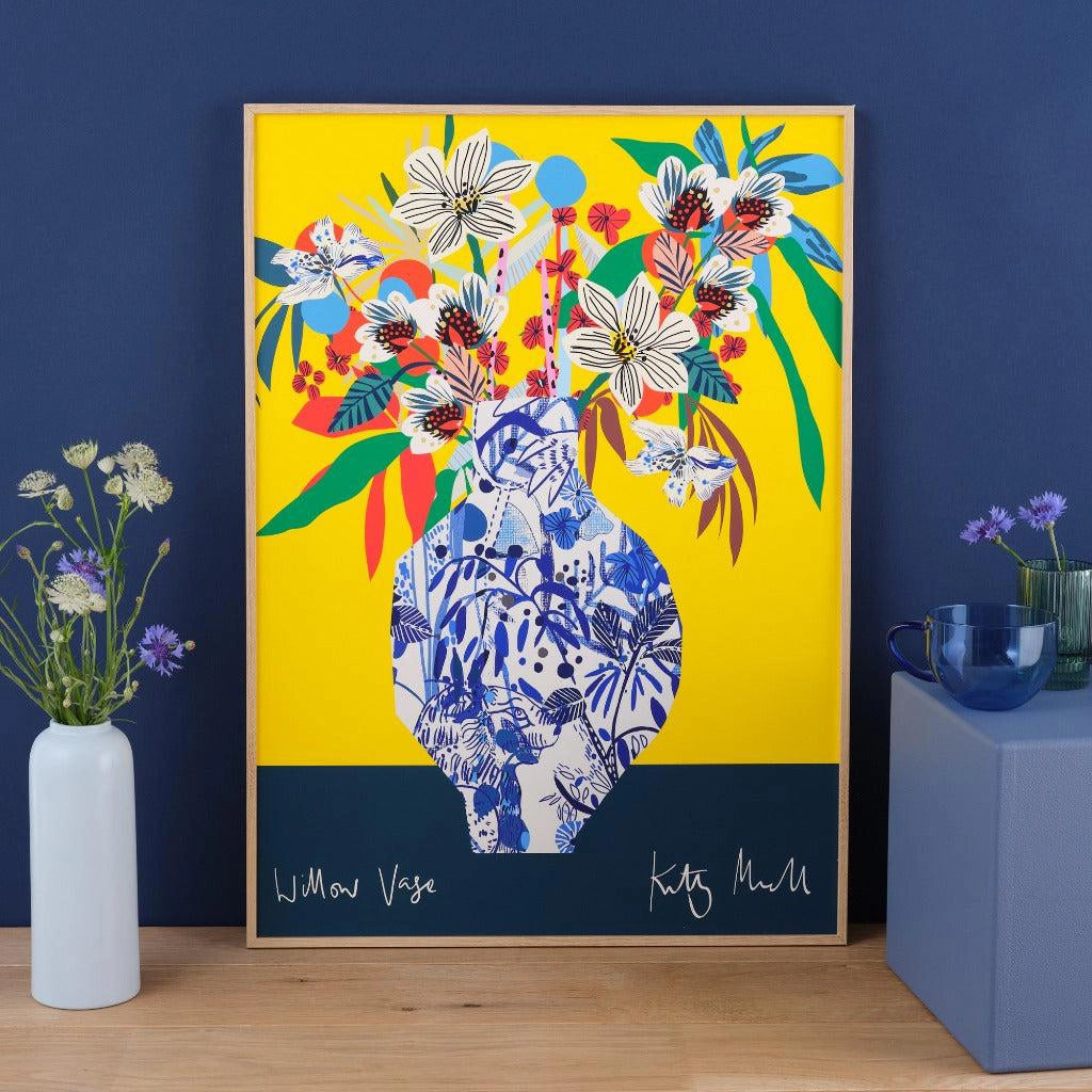 Willow Vase fine art print by Kitty McCall in a natural oak frame, styled on a wooden surface alongside wildflowers and a blue mug against a navy wall